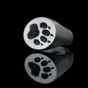 Könnte beinhalten: Ein silberner Ring mit einem Bärenpfotenabdruck-Design. Der Ring ist oval geformt und hat eine glatte, polierte Oberfläche.