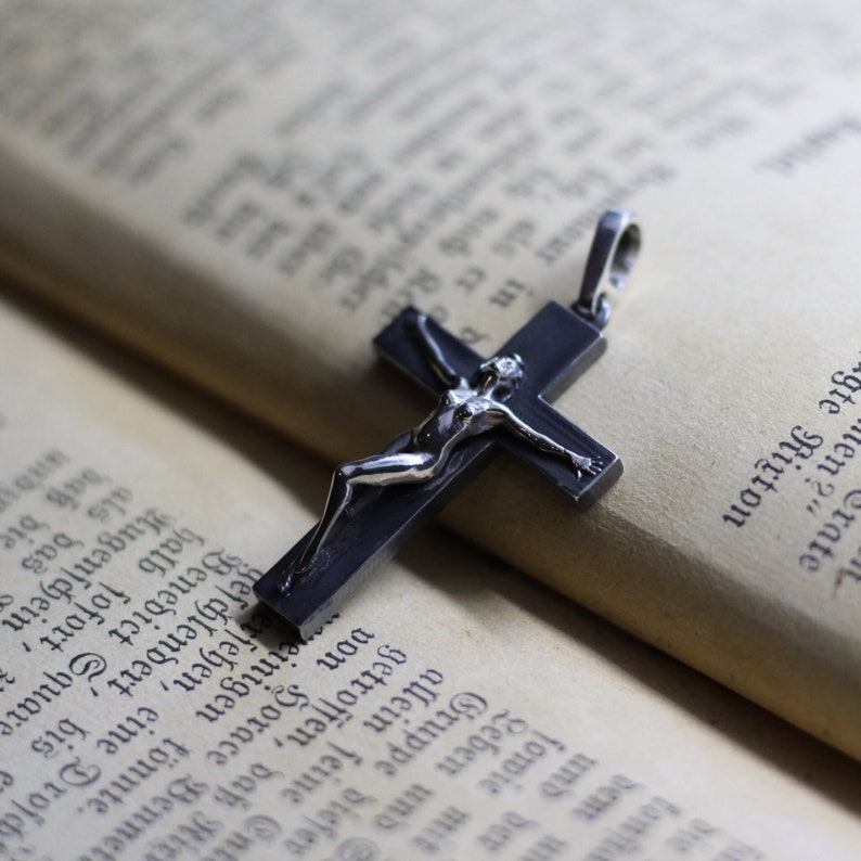 May include: A dark metallic cross pendant featuring a polished silver-toned figure attached to the front. The cross has a smooth, matte finish and includes a small metal loop at the top for attachment to a chain or cord, presented as a jewelry item.