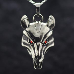 Puede incluir: Un collar con colgante plateado que presenta una cabeza de rata con ojos rojos. La cabeza de la rata está detallada con orejas visibles, un hocico puntiagudo y una superficie texturizada. El colgante cuelga de una cadena plateada, sobre un fondo oscuro.
