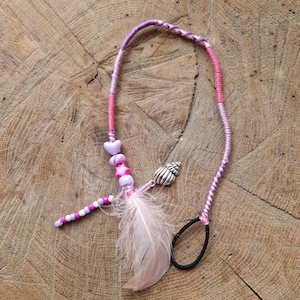 May include: A handmade hair wrap featuring a pink and purple braided cord. Adorned with heart-shaped beads, a seashell charm, a pink feather, and a beaded accent. The hair wrap is secured with a black elastic band.