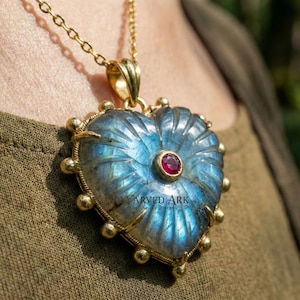 Puede incluir: Un collar dorado con un colgante en forma de corazón. El colgante presenta una piedra tallada azul-verdosa con una pequeña gema roja en el centro. El colgante está rodeado de detalles dorados y una cadena dorada.