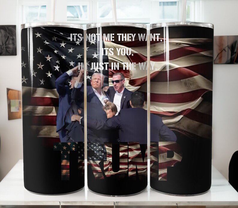 Może przedstawiać: Trzy kubki ze stali nierdzewnej z czarnym tłem i wzorem przedstawiającym flagę amerykańską, grupę ludzi i tekst "ITS NOT ME THEY WANT. ITS YOU. IM JUST IN THE WAY." Tekst "TRUMP" jest wyświetlany dużymi literami z wzorem amerykańskiej flagi wewnątrz liter.