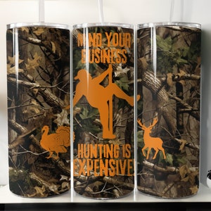 Op de afbeelding: Drie roestvrijstalen bekers met een camouflagepatroon. De middelste beker heeft de tekst "Mind Your Business Hunting Is Expensive" in oranje. De linker beker heeft een kalkoensilhouet en de rechter beker heeft een hertensilhouet.