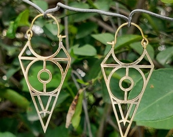 Peut inclure: Une paire de boucles d'oreilles créoles géométriques dorées en forme de triangle avec un motif circulaire au centre.