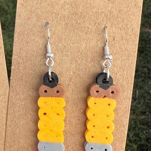 Puede incluir: Un par de pendientes colgantes hechos de arcilla polimérica. Los pendientes tienen forma de lápices y son amarillos, marrones, grises y rosas. Los pendientes cuelgan de ganchos plateados.