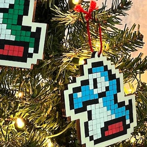 May include: Pixelated Christmas ornaments featuring video game characters. One ornament depicts a blue and white character with a red tongue, hanging from a red string. The ornaments are displayed on a Christmas tree with lights.
