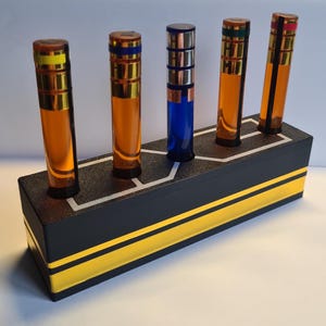 May include: A black wooden stand with gold stripes holds five small glass bottles with coloured caps. The bottles contain liquid and are arranged in a row.