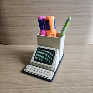 May include: A retro-style desk organizer shaped like a computer. It features a screen with green vertical lines, a keyboard, and a pen holder containing pink and orange highlighters and a green pen. The organizer is made of light gray plastic.