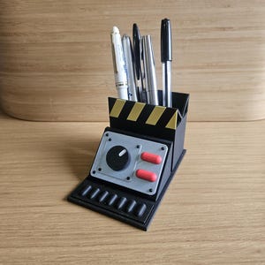 May include: A black desk tidy with a control panel design, holding pens. The tidy features a gray panel with a dial and two red buttons. The base has parallel lines. The pens are of various colours.