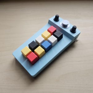 Könnte beinhalten: Ein hellblaues, rechteckiges Makro-Keypad mit einem Raster aus zehn quadratischen Tasten in Rot, Gelb, Weiß, Schwarz und Blau. Das Gerät verfügt über zwei kleine schwarze Drehregler und einen größeren silbernen runden Knopf an der Oberseite.