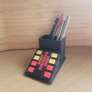 May include: Black desk organizer with a graphic equalizer design in red and yellow. The organizer holds pens and pencils. The base has a series of red and yellow squares. The organizer is on a wooden surface.