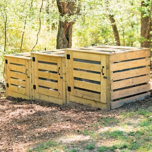 Op de afbeelding: Een houten compostbak met drie secties, opgebouwd uit horizontale lichtbruine houten latten met zwarte metalen scharnieren, grendels en handgrepen op elke deur. De structuur is voorzien van gaaspanelen tussen de latten voor ventilatie.