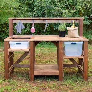 Peut inclure: Un établi de jardinage en bois brun doté d'une étagère supérieure avec cinq crochets noirs, d'un plan de travail central, d'une étagère inférieure et de deux bacs en plastique transparent. Un chiffon gris, une brosse rose, des ciseaux, une plante en pot et un sac en toile de jute sont inclus.