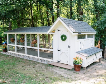 May include: A white wooden chicken coop with a gray shingled roof, featuring a long wire mesh run, a main door with a green wreath, and a side nesting box. Hanging flower baskets are attached to the run, and potted flowers sit nearby.