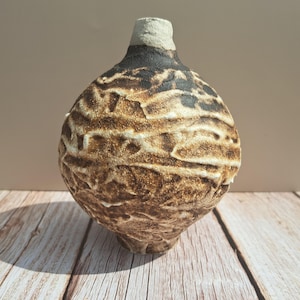 May include: A handmade ceramic vase with a textured, layered design. The vase has a rounded body and a slender neck. The colour scheme includes shades of brown, tan, and black, creating a rustic aesthetic. The vase is suitable for home decor.