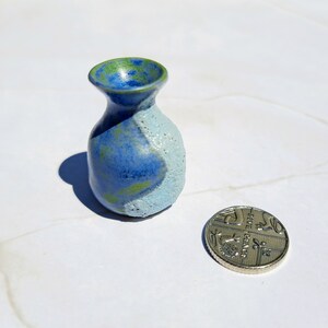 Pode incluir: Um pequeno vaso de cerâmica azul e verde com um esmalte branco de um lado. O vaso é aproximadamente do tamanho de uma moeda britânica de uma libra.