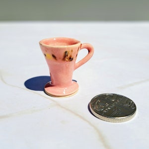 May include: A small pink ceramic cup with a brown and yellow design. The cup has a flared base and a handle.