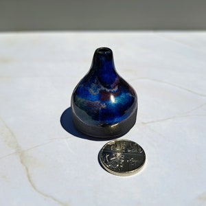 Pode incluir: Um pequeno vaso de cerâmica azul escuro com detalhes iridescentes azuis, roxos e dourados. O vaso tem um corpo arredondado e um pescoço estreito.