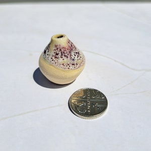 Pode incluir: Um pequeno vaso de cerâmica em forma de pêra com um esmalte amarelo e manchas roxas. O vaso é aproximadamente do tamanho de uma moeda britânica de uma libra.