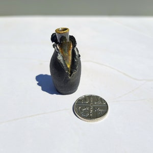 Op de afbeelding: Een kleine, handgemaakte keramische vaas met een donkere, getextureerde buitenkant en een goudkleurige binnenkant. De vaas heeft een unieke, organische vorm en een smalle opening. Een munt ligt ernaast ter vergelijking.