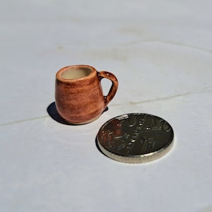 Pode incluir: Uma pequena caneca de cerâmica marrom com alça. A caneca é do tamanho de uma moeda de 1 euro.