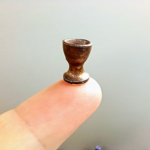 Pode incluir: Uma pequena xícara de cerâmica dourada com uma base arredondada. A xícara está sentada em um dedo.