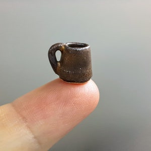 Pode incluir: Uma pequena caneca de cerâmica marrom com alça. A caneca está apoiada em um dedo.
