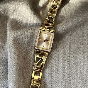 Può includere: Orologio Guess dorato con quadrante rettangolare e lunetta tempestata di diamanti. L'orologio presenta un cinturino dorato con un design geometrico aperto. Il quadrante è di colore champagne chiaro.