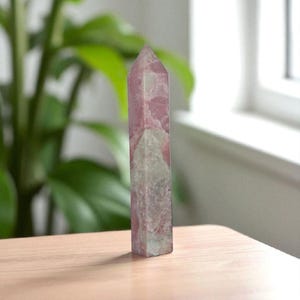 May include: A tall, pink and white crystal point stands on a light brown surface. The crystal has a rectangular base and tapers to a sharp point. The colors are a mix of pink and white, with some areas appearing translucent. The background includes green foliage and a window.