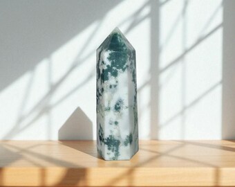 May include: A tall, polished crystal obelisk with a pointed top. The crystal is primarily white with green, moss-like inclusions. It stands on a wooden surface, with shadows from a window casting across the background.