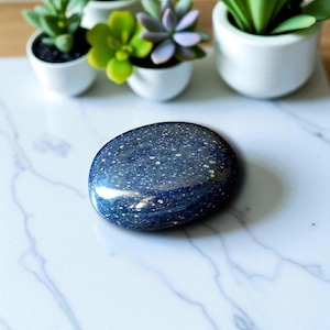 May include: A polished, oval-shaped stone with a deep blue hue and speckled with small, light-colored inclusions. The stone has a glossy finish and reflects light. The background includes small potted succulents on a white marble surface.