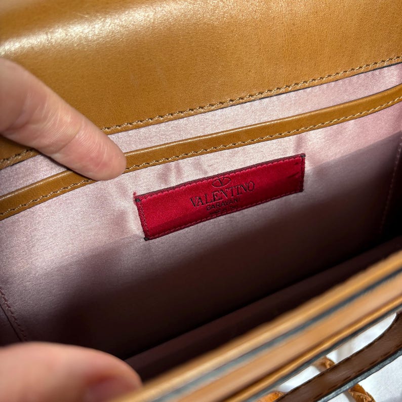 May include: Tan leather handbag interior with a pink satin lining. A red rectangular label with the brand name "VALENTINO GARAVANI" is sewn into the lining. The bag's interior is visible.