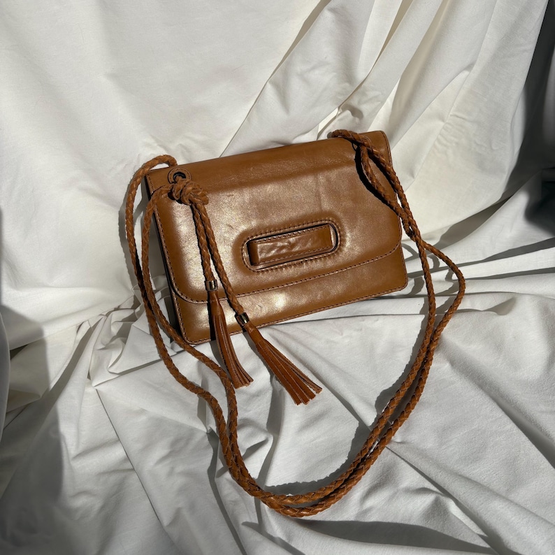 May include: A brown leather crossbody bag with a rectangular shape and a flap closure. The bag features a braided strap and decorative tassels. The bag is approximately 9 inches wide and 6 inches tall.
