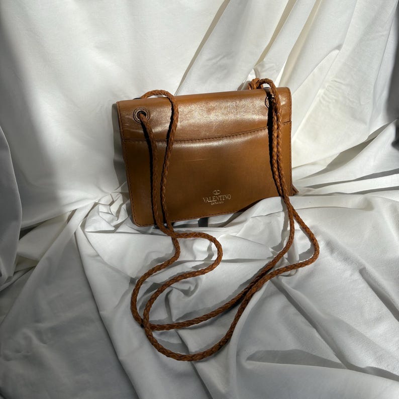 May include: A brown leather crossbody bag with a braided strap. The bag has a rectangular shape with rounded corners and a flap closure. The Valentino logo is visible on the front of the bag.