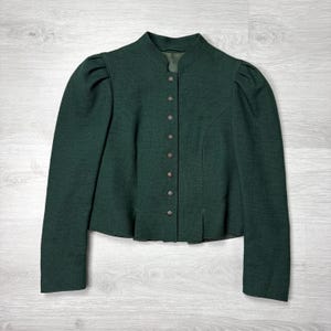 May include: A dark green jacket with a stand-up collar and long sleeves. The jacket has a row of buttons down the front and subtle pleats at the waist. The fabric appears to be a textured wool or wool blend.