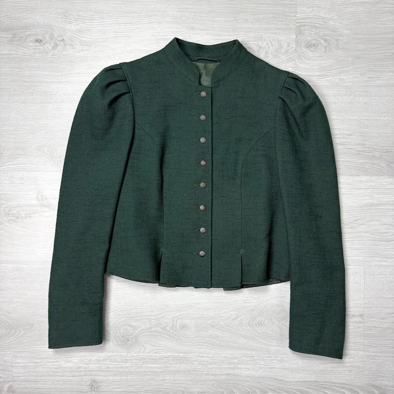 May include: A dark green jacket with a stand-up collar and long sleeves. The jacket has a row of buttons down the front and subtle pleats at the waist. The fabric appears to be a textured wool or wool blend.