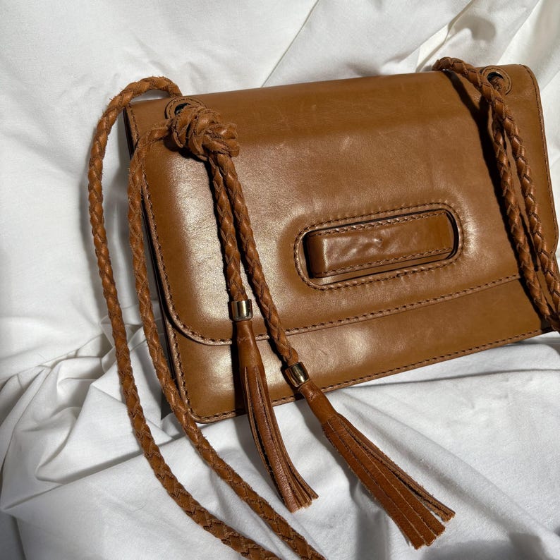 May include: A brown leather handbag with a flap closure and a braided shoulder strap. The bag features a rectangular design with a decorative handle and two tassels. The bag is set against a white background.