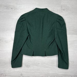 May include: A dark green blazer with a tailored fit, featuring a stand-up collar and structured shoulders. The back view showcases a classic design with long sleeves and a subtle pleat detail at the hem. The jacket is made of a textured fabric.