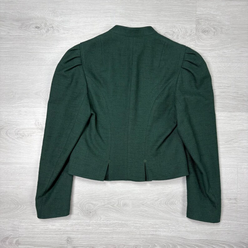 May include: A dark green blazer with a tailored fit, featuring a stand-up collar and structured shoulders. The back view showcases a classic design with long sleeves and a subtle pleat detail at the hem. The jacket is made of a textured fabric.