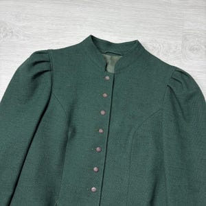 May include: A dark green jacket with a high collar and button-down front. The jacket has long sleeves with slight puffing at the shoulders. The buttons are small and round, and the fabric has a subtle texture.
