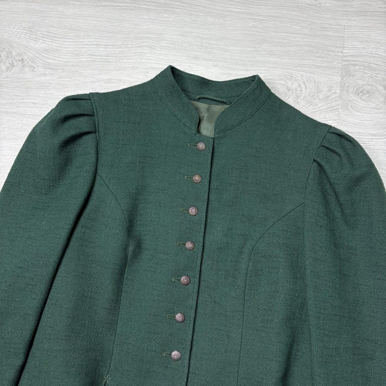 May include: A dark green jacket with a high collar and button-down front. The jacket has long sleeves with slight puffing at the shoulders. The buttons are small and round, and the fabric has a subtle texture.