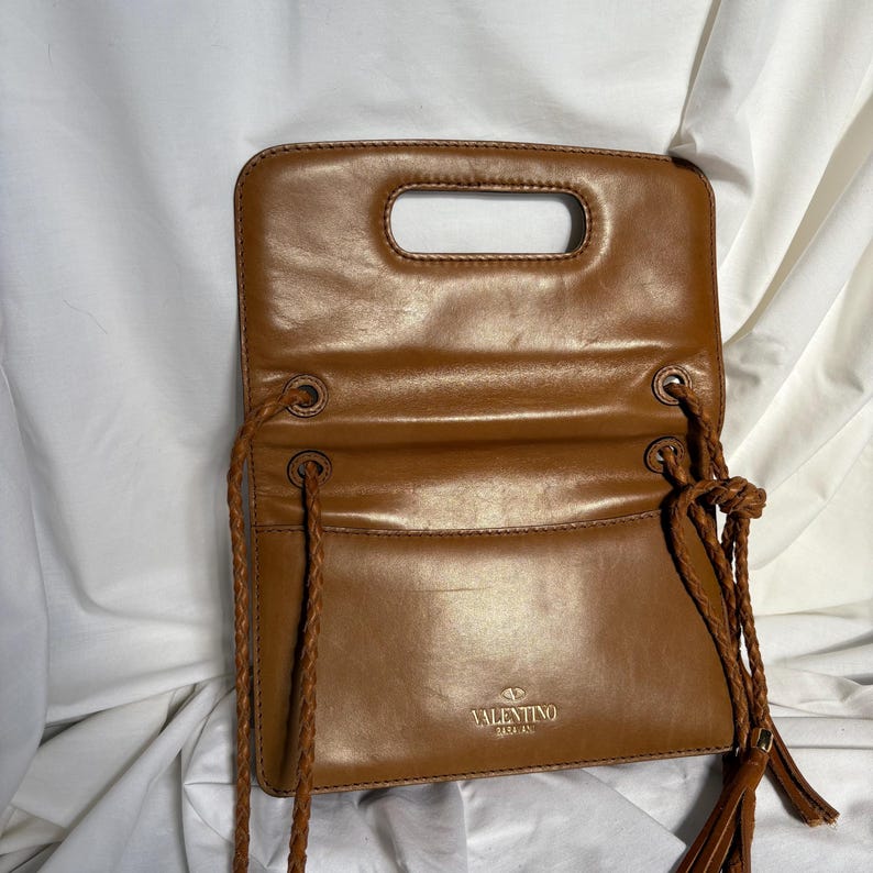 May include: A brown leather handbag with a handle opening at the top. The bag has a flap closure and braided rope straps with tassels. The Valentino logo is visible on the front of the bag.
