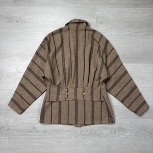 May include: A brown and black striped jacket with a collar and a belt detail. The jacket has long sleeves and two gold-colored buttons on the belt. The jacket is made of a textured fabric.