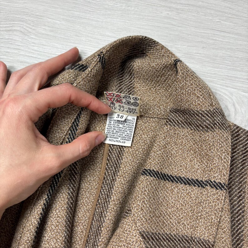 May include: Close-up of a tan and brown patterned jacket. The jacket has a label with washing instructions and the number 38. The jacket is held by a person's hand.