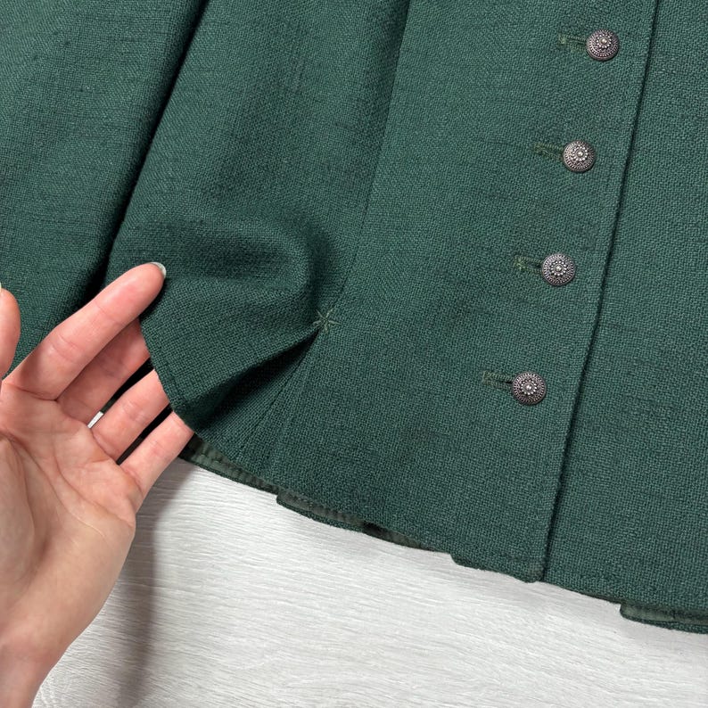 May include: Close-up of a dark green jacket with four decorative buttons. The jacket's fabric has a subtle texture, and the buttons are detailed with a circular design. The jacket's edge is visible, showing the tailored construction.