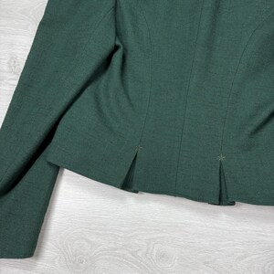 May include: Close-up of a dark green blazer. The blazer features a tailored design with a notched lapel and a two-button closure. The fabric has a subtle texture, and the sleeves are long. The back of the blazer has pleats.