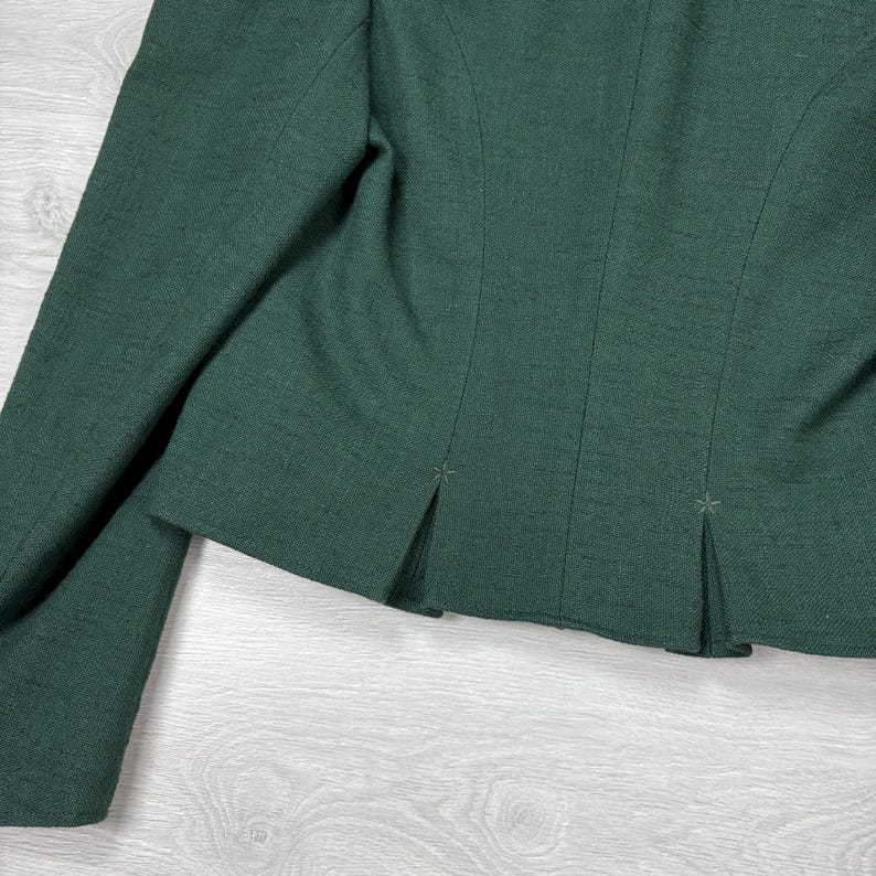 May include: Close-up of a dark green blazer. The blazer features a tailored design with a notched lapel and a two-button closure. The fabric has a subtle texture, and the sleeves are long. The back of the blazer has pleats.