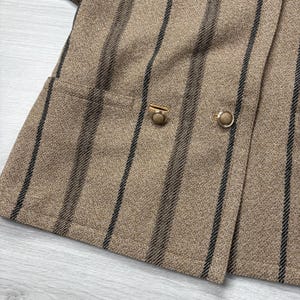 May include: Close-up of a tan blazer with vertical black stripes. The blazer features a double-breasted design with two gold-colored buttons and a small pocket. The fabric has a textured appearance.