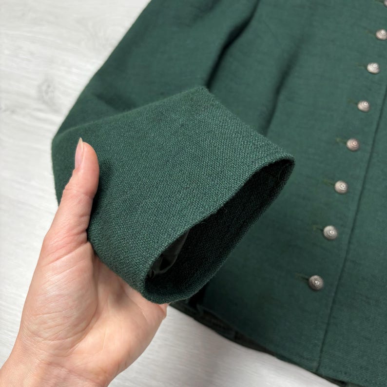 May include: Close-up of a dark green jacket sleeve. The sleeve is made of a textured fabric and is cuffed. Silver buttons line the front of the jacket. The jacket appears to be a traditional style.