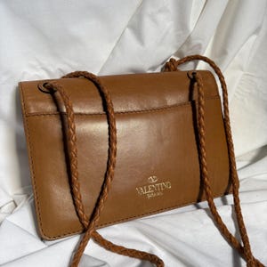 May include: A brown leather crossbody bag with a braided strap. The bag has a rectangular shape with a flap closure and a small pocket on the back. The Valentino Garavani logo is visible on the front.